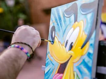 EPCOT International Festival of the Arts Man painting on canvas and the painting is of Pluto the dog, a yellow cartoon dog, with a blue background.