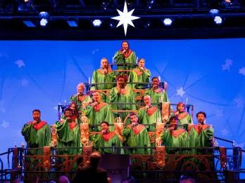 EPCOT International Festival of the Holidays Choir stood on steps which makes them form the shape of a Christmas party and they are wearing green capes with red collars. There is a star at the top.