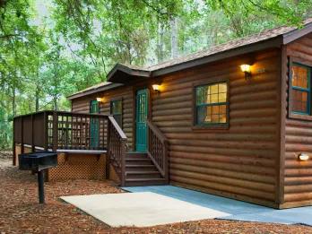 small, clean wooden cabin in the woods with a small black grill next to it.
