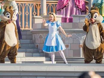 Mickey’s Magical Friendship Faire Alice from Alice in Wonderland stood on stage waving at guests, wearing her iconic blue dress with a white apron over, black bow in her hair, and white tights. Next to her either side are the two cartoon chipmunks, one with a black nose, the other with a red nose.
