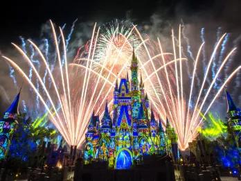 Happily Ever After Large pink and blue castle with projections of Disney characters on including Cinderella, a blonde woman with a blue ballgown on. There are fireworks behind and surrounding the castle lighting up the night sky.