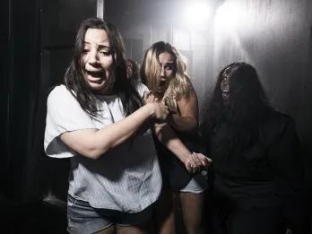 Two young women in a dark room with a bright light shining behind with terrified facial expressions, clutching onto one another and dodging another person who is lurking in the dark. The third person has black hair and a wound on their head which seems to be bleeding down their face. Their hair is messy and they are pulling a scary face with their tongue poking out.