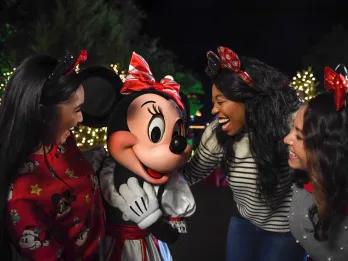 Mickey’s Very Merry Christmas Party Minnie mouse stood in a white and red stripey bow and a matching dress. There are three women crowded around her, one on the left wearing a red jumper with festive Disney cartoon characters on and the other two on the right wearing jumpers and jeans and sparkly Minnie mouse ear headbands.