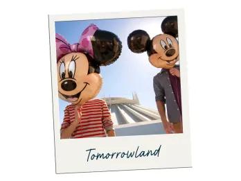 Tomorrowland Photo of short person with red and white t-shirt on holding a Minnie Mouse balloon in front of their head. To the right there is a larger person holding a mickey mouse balloon in front of their head. In the background there is a large white building shaped like a spaceship.