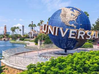 Large circular sign shaped as the globe with white letters in bold curving around it spelling out "UNIVERSAL". In the background there is a lake, palm trees and a tall sign made from rock which says Islands of Adventure.