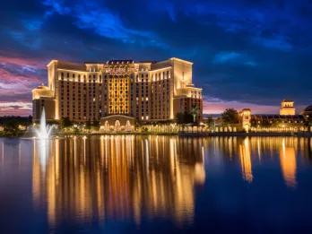 Large clean building which is cream coloured and lit up in yellow lighting in the night time. There is a large lake with a fountain sprinkling water up.