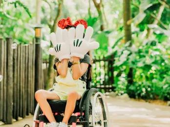 Discovery Island Trails Young child in a wheelchair covering their face with two hands which have large white mickey gloves on. They are wearing red sparkly mickey ears on their head. In the background there is a pathway with trees surrounding it.