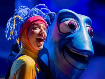 Finding Nemo: The Big Blue… and Beyond A woman with a yellow full body suit which turns red on her head. On the top of her head she has clue strands coming off which looks like pieces of a coral reef. To the right is a large puppet of Dory, a blue fish staring in the same direction as her.