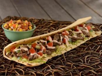 Long taco with beef, tomatoes, salad and a white sauce zig zagging throughout. Next to it looks like a small bowl of rice.