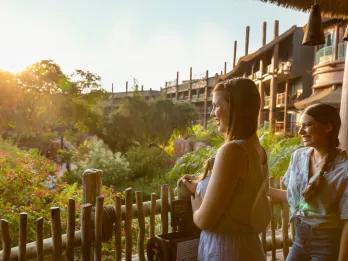 Two young women stood on a balcony with a wooden railing in a brown wooden building overlooking gorgeous gardens and greenery.