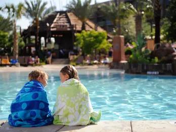 Two young girls sat in towels beside the pool, dangling their legs into the pool. In the background there is a brown shack and trees. There are also lounge chairs.