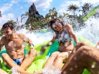 Volcano Bay Four young adults sat on a green rubber ring splashing into a large pool with a large statue of a volcano in the background.