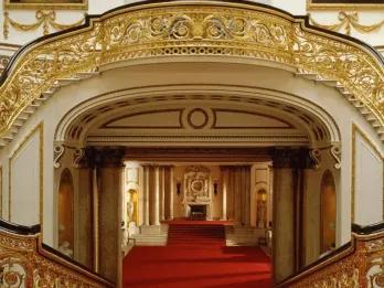 Buckingham Palace staircase Golden and wooden bannisters lining two staircases which wrap around and join to create on large staircase. There is bright red carpet and white walls lined with gold details. There is a large open hallway with a fireplace at the end.
