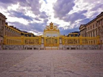 Golden gates with beautiful royal details, there are stone statues and behind is a grand palace.