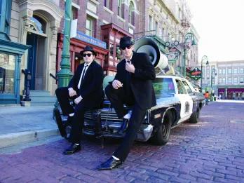 Blues Brothers Two men in black suits and hats are leaning against a black and white car which has a large speaker on top of it. They are in the middle of the New York set streets.