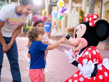 Minnie Mouse Meet and Greet Large cartoon mouse wearing a red dress with white polka dots on it and a matching bow. There is a young boy touching Minnie Mouse's nose. There is a young girl next to the boy and a man bending down smiling and watching. In the background there are multi-coloured Mickey balloons.