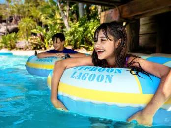 Castaway Creek Little girl and little boy smiling inside of rubber rings on a lazy river. They are floating under a bridge and there are many trees behind them.