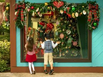 Christmas Tree Trail Two kids, one boy and one girl, stood in shop window looking inside wearing Mickey Mouse ear shaped hats. Inside there is a Christmas tree lavishly decorated and above the window there are christmas decorations.