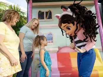 Gabby Meet and Greet Large child cartoon character with black hair in pigtails and an animal headband looking at a young girl meeting her. There are also two women stood behind watching and smiling.