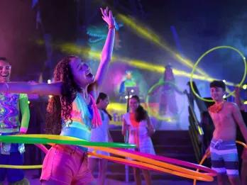 H2O Glow Girl laughing in neon coloured glowing swimsuit hula hooping with multiple hoops. She has one arm up in the air. Around her there are multiple people watching and smiling.