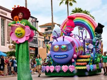 Mega Movie Parade Woman on stilts dressed like a flower with a face on it with a huge hat on. Behind her pink troll called Poppy riding a caterpillar creature on a float is waving at crowds.