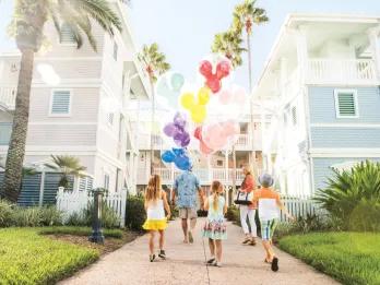 Family of 5 guests walking between the tall pale blue and white aesthetic buildings holding Mickey Mouse shaped balloons of different colours.