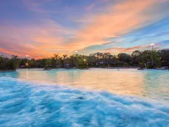 Open Surf Sun set with a blue sky and pink/orange coloured clouds. There is a huge pool with a wave splashing through which is surrounded by trees.