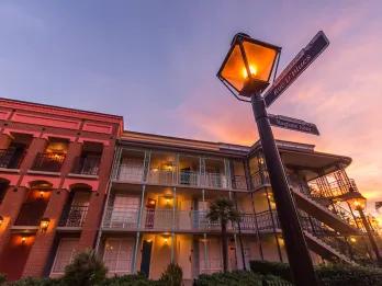Old fashioned buildings coloured orange, red and pale yellow glowing in the sunlight reflecting from the sunset in the background. There is an old-fashioned lamp pot with wooden signs pointing different directions. 