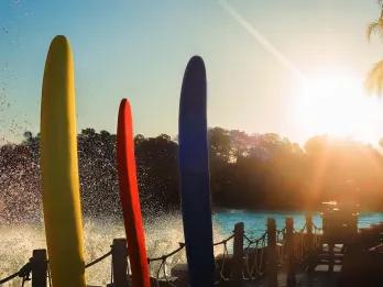 Private Surf Three multicoloured surf boards stood up with wave hitting the side behind them and a large pool as the sun rises.