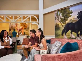 Two guests sat on a sofa looking towards a sat cast member who is smiling telling them information. The lobby is beige with large paintings of cartoon horses on the walls.