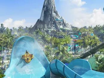 Honu, Volcano Bay Four people facing each other on a large yellow rubber ring flying up a light blue water slide wall which is huge. In the background, there is a volcano statue with slides coming off of it and water.