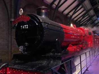 Hogwarts Express Large steam train with the label Hogwarts Express, coloured black and a dark red. There is steam surrounding and it is parked in a large factory type building.