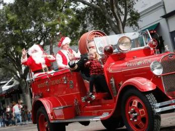 An old fashioned firetruck in middle of a road with man dressed as santa claus sat in the back waving at people. There is also someone sat in the front with a little girl.