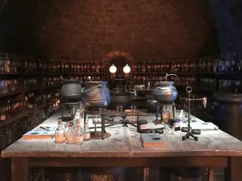 A dark lit room with a dimly lit main table with multiple black cauldrons and substances. In the background there are dark shelves holding test tubes with different potions and substances.