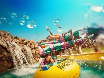 In the background there are many large waterslides wrapping around one another in different colours. There is a small lazy river flowing with a waterfall pouring into it off of rocks. There is a mother holding her little girl who is sat on a yellow rubber ring. There are fountains also splashing them.
