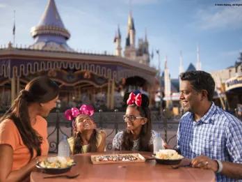 Dining Plan Family of four dining together in Magic Kingdom Park with carousel behind them. The two little girls are wearing minnie ears and smiling.