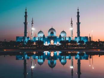 Abu Dhabi A beautiful religious temple in Abu Dhabi located overlooking a lake. The sun is setting with a pink and blue hue in the sky and the temple is perfectly reflected in the lake. It is a white building with three white domes and four big pillars sticking out.