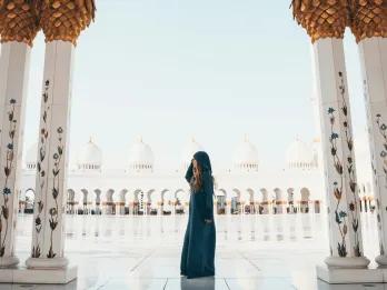 Abu Dhabi Sightseeing There is a gorgeous sleek tiled floor which is so clean you can see a reflection of people in it. There's a woman wearing a robe which covers her whole body to be respectful to the culture. There are pillars which are white and gold with paintings of flowers growing up them and on the other side it is the same, with white domes on top of the pillars.