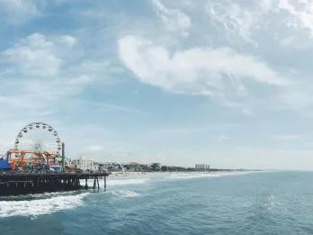 There is the sea waves crashing onto a yellow sandy beach. There is also a long iconic pier with a funfair on it, including a Ferris wheel and small rollercoaster. In the far distance there are only a few large buildings..
