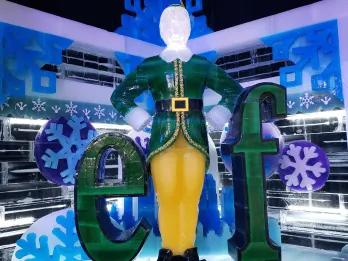Sign of green letters saying elf in font from movie but instead of an L there is an ice sculpture of Buddy the Elf stood up straight wearing a dark green elf suit.