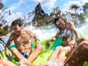 Volcano Bay 4 people sat in a big rubber ring heading into a large pool of water which is splashing them as they cling on. In the background there is a large brown/grey coloured volcano.