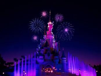 Sleeping Beauty Castle Castle lit neon pink in the night sky with purple fountains below. There are also pink fireworks in the sky.