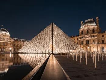 A glass panelled pyramid building in the centre of old fashioned castle like buildings. It is reflecting in the small pond.