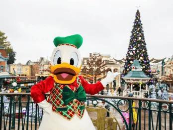 Donald Duck standing on a balcony pointing towards a large Christmas tree in the distance