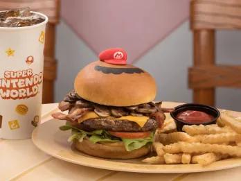 Ein großer Burger mit einem Brioche-Brötchen, einem großen Rindpatty, Käse, Champignons, Salat, Tomate und einer kleinen roten Mario-Mütze darauf. Dazu gibt es Pommes und einen kleinen schwarzen Topf mit Ketchup. Außerdem gibt es einen großen Becher Cola.