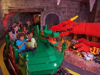 Kids and parents on rollercoaster, the car is a lego green dragon and it is going through a room with a large red dragon inside guarding jewels in a castle.