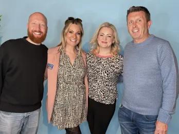 Four employees stood in front of a blue screen smiling. One has the do something different badge on her blazer.