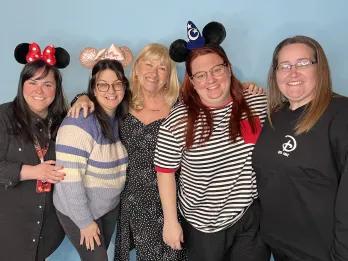 Five women employees wearing Disney ears and a Disney jumpers hugging in front of a blue screen.
