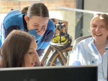 Marketing Three girls gathered around a computer laughing, with one covering her mouth with her jumper