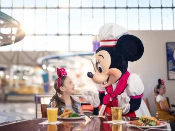 Minnie Mouse dressed in a chef hat and costume meeting a girl with pink sparkly mickey mouse headbands. There are different dishes on the table from the buffet.